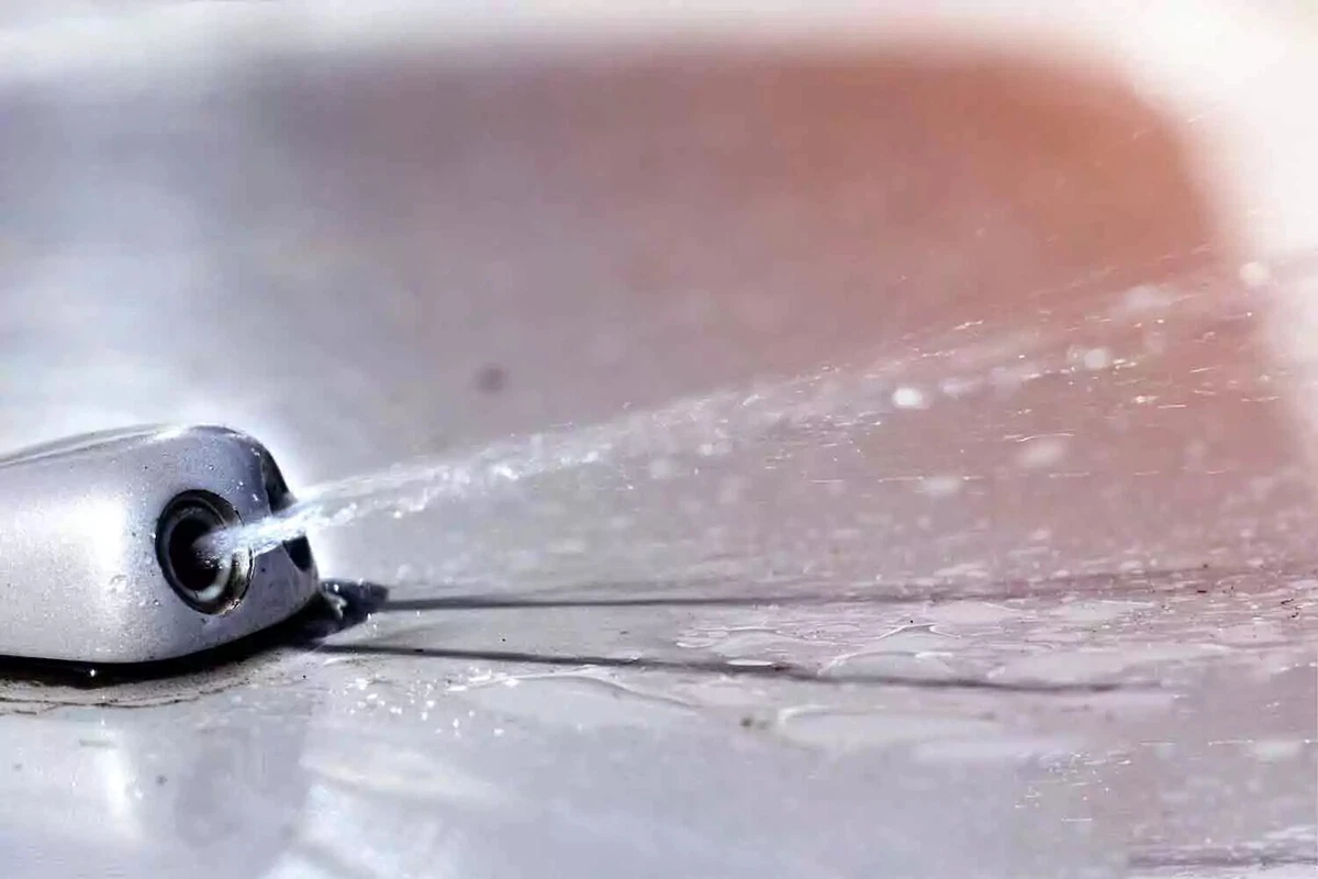A close-up view of a silver car’s windshield washer nozzle spraying fluid.