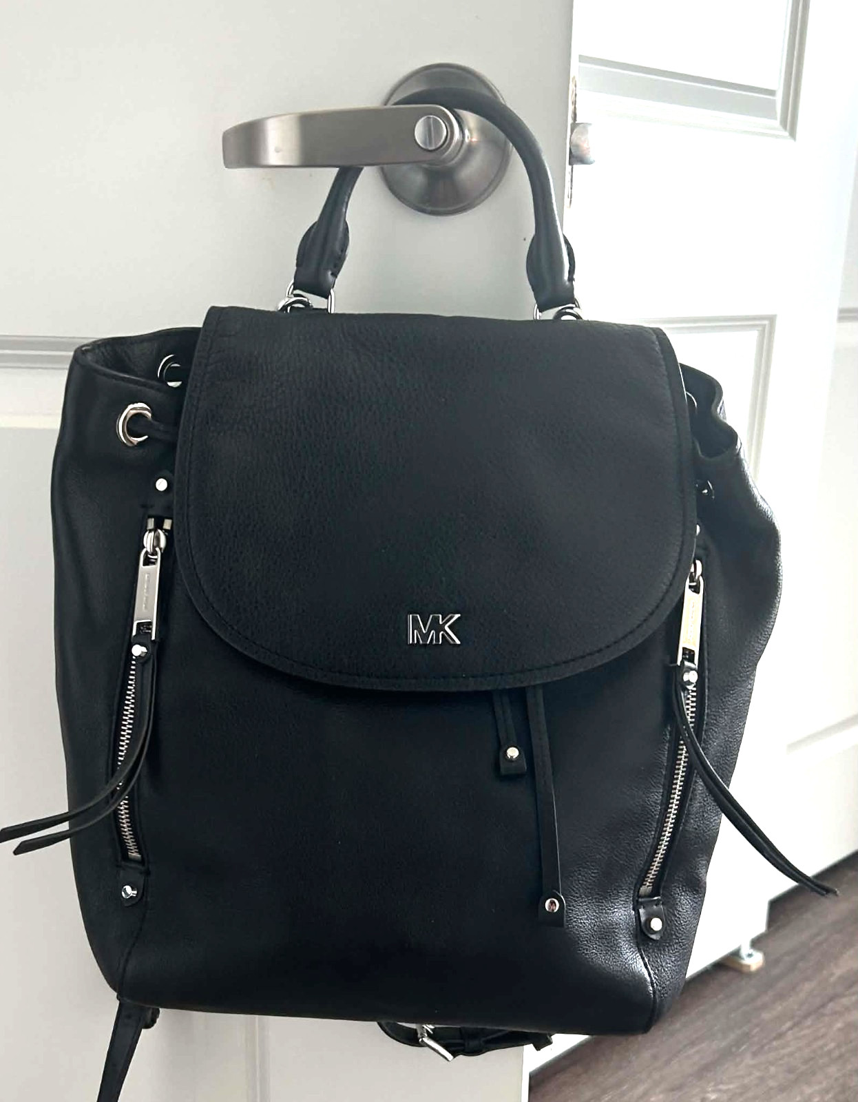 Black pebbled leather Michael Kors Evie backpack with a rounded flap top, hanging from a door handle.