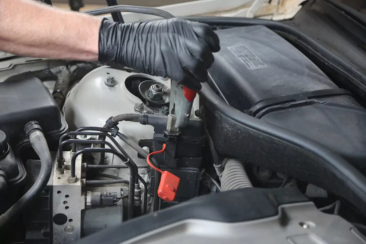 A mechanic wearing a black glove places the red handled jumper cable on the battery's positive post.