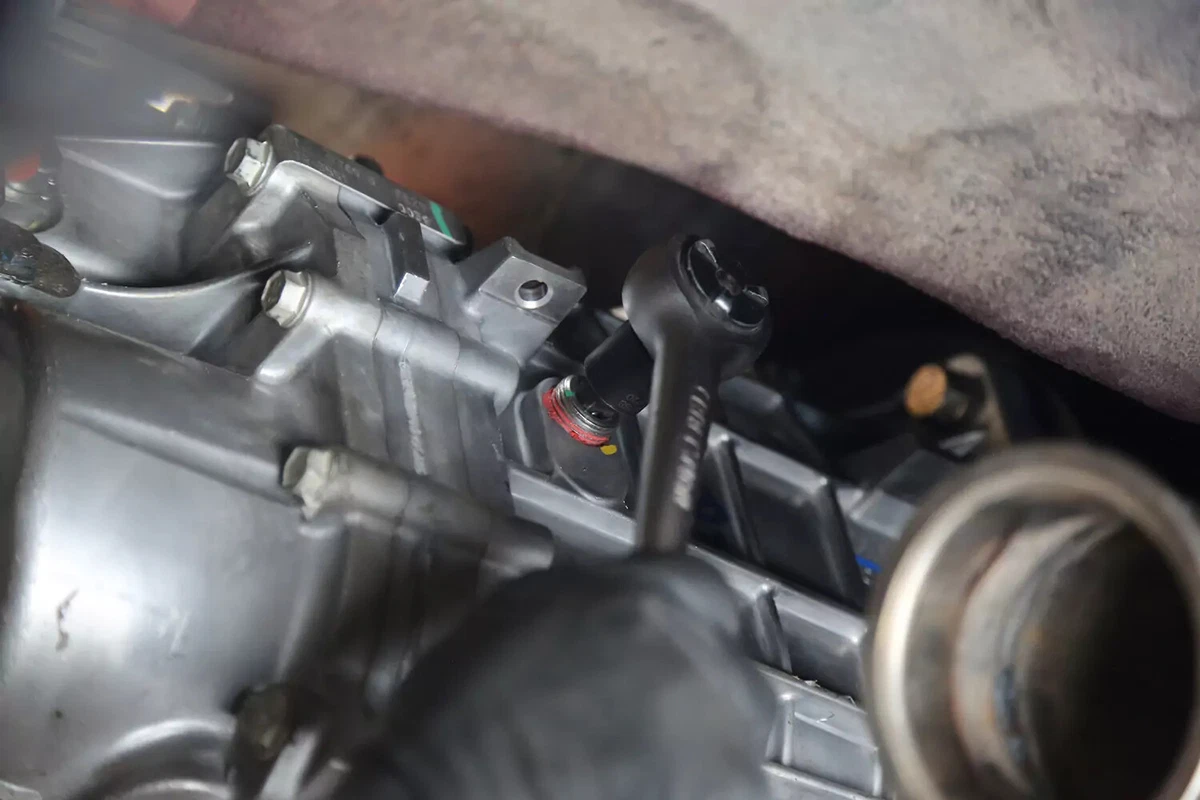 A mechanic wearing a black glove uses a socket wrench to remove the fill plug of a manual transmission.