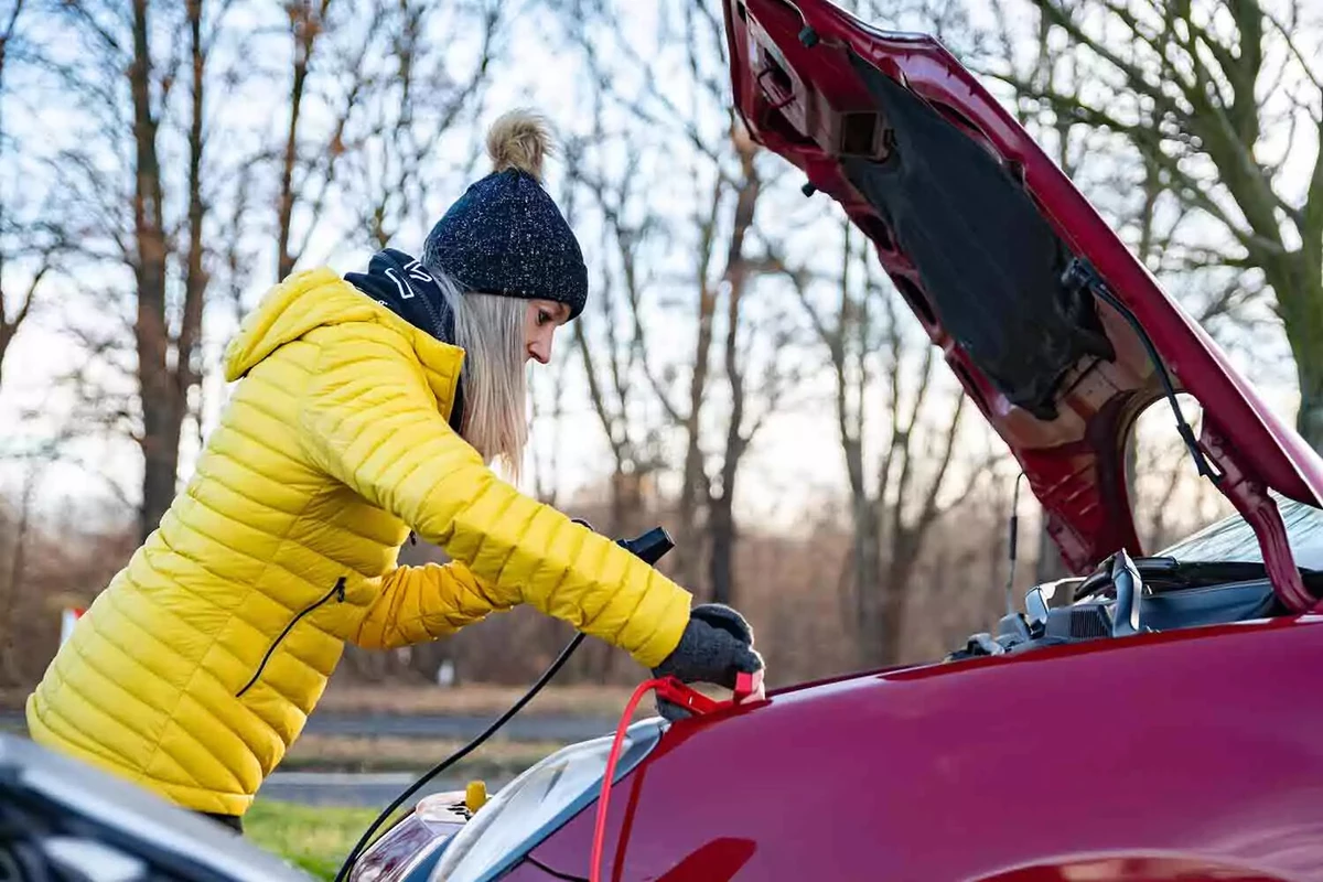 A woman in a puffy yellow winter coat connects jumper cables to the battery in a red car.