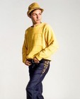 Man wearing yellow knit cardigan with black pants, brown jacket, and tan hat on light gray background.