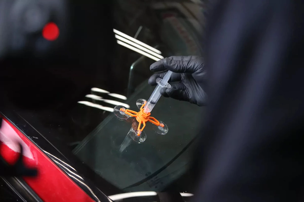 A mechanic wearing a black glove depresses the plunger of the syringe in the repair jig of a windshield repair kit.