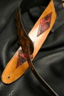 Black leather guitar strap with red and silver inlay featuring diamond patterns, displayed on a textured black surface.