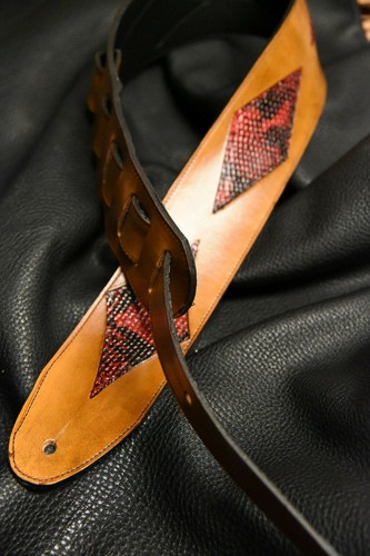 Black leather guitar strap with red and silver inlay featuring diamond patterns, displayed on a textured black surface.