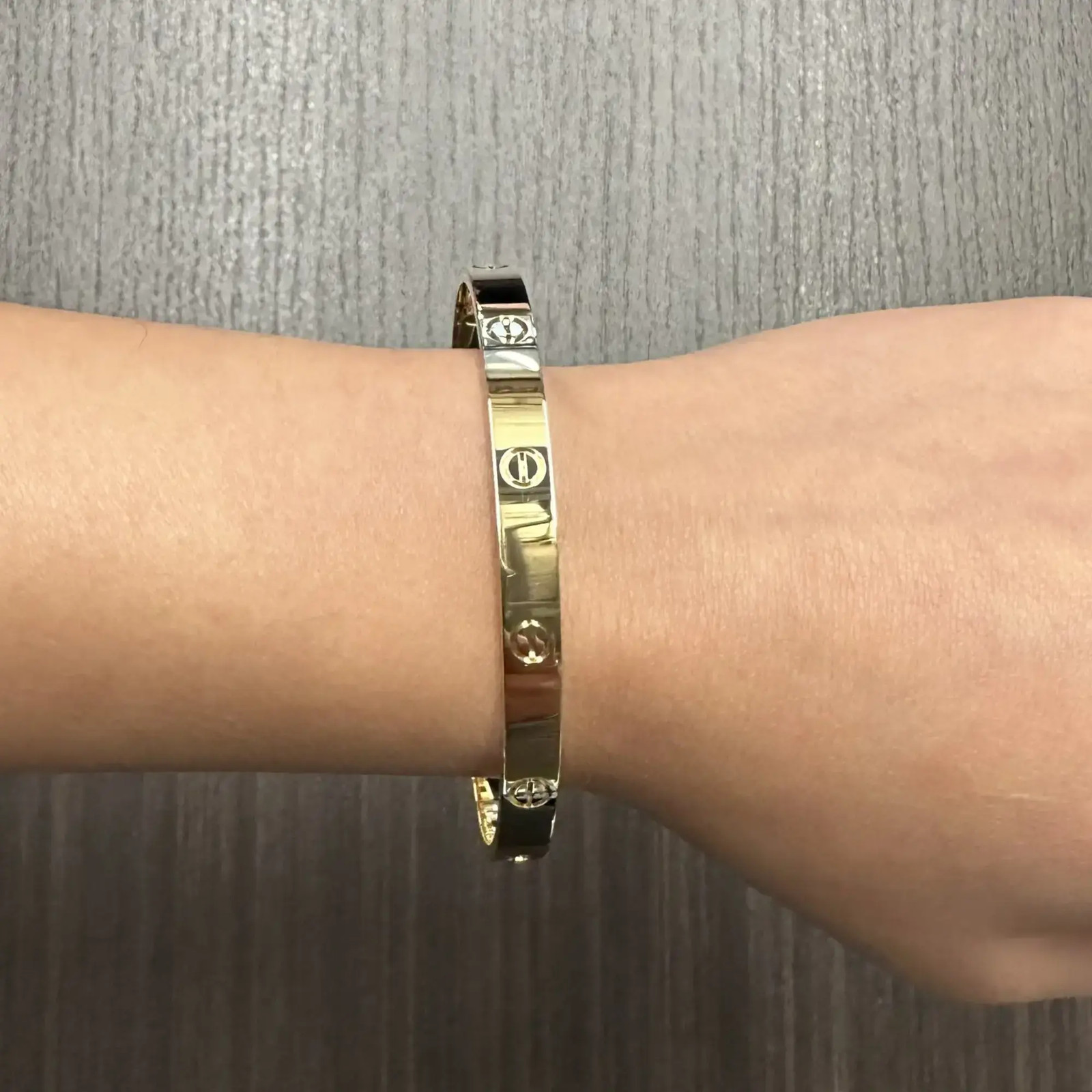 An image of a unisex adult Cartier love bracelet without stones, displayed on a wrist at a close-up angle. The bracelet is centered in the frame, showing a front-facing view with a slight tilt towards the camera, allowing for a clear view of its design and clasp detail. The background is a blurred texture of a grey surface, emphasizing the bracelet on the forearm. Great preowned condition.
