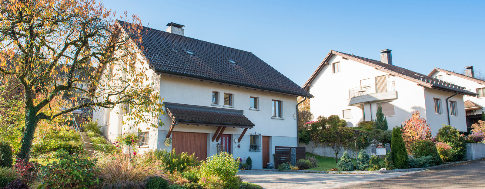 Ein modernes Wohnhaus mit weißer Fassade und dunklem Schrägdach in einer ruhigen Nachbarschaft. Vor dem Haus befindet sich ein gepflegter Garten mit bunten Pflanzen und einem großen Baum, der herbstlich gefärbte Blätter trägt.