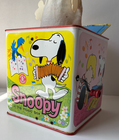 White Snoopy tin lunchbox with red band, blue cap, and text “In the music box” on green background.