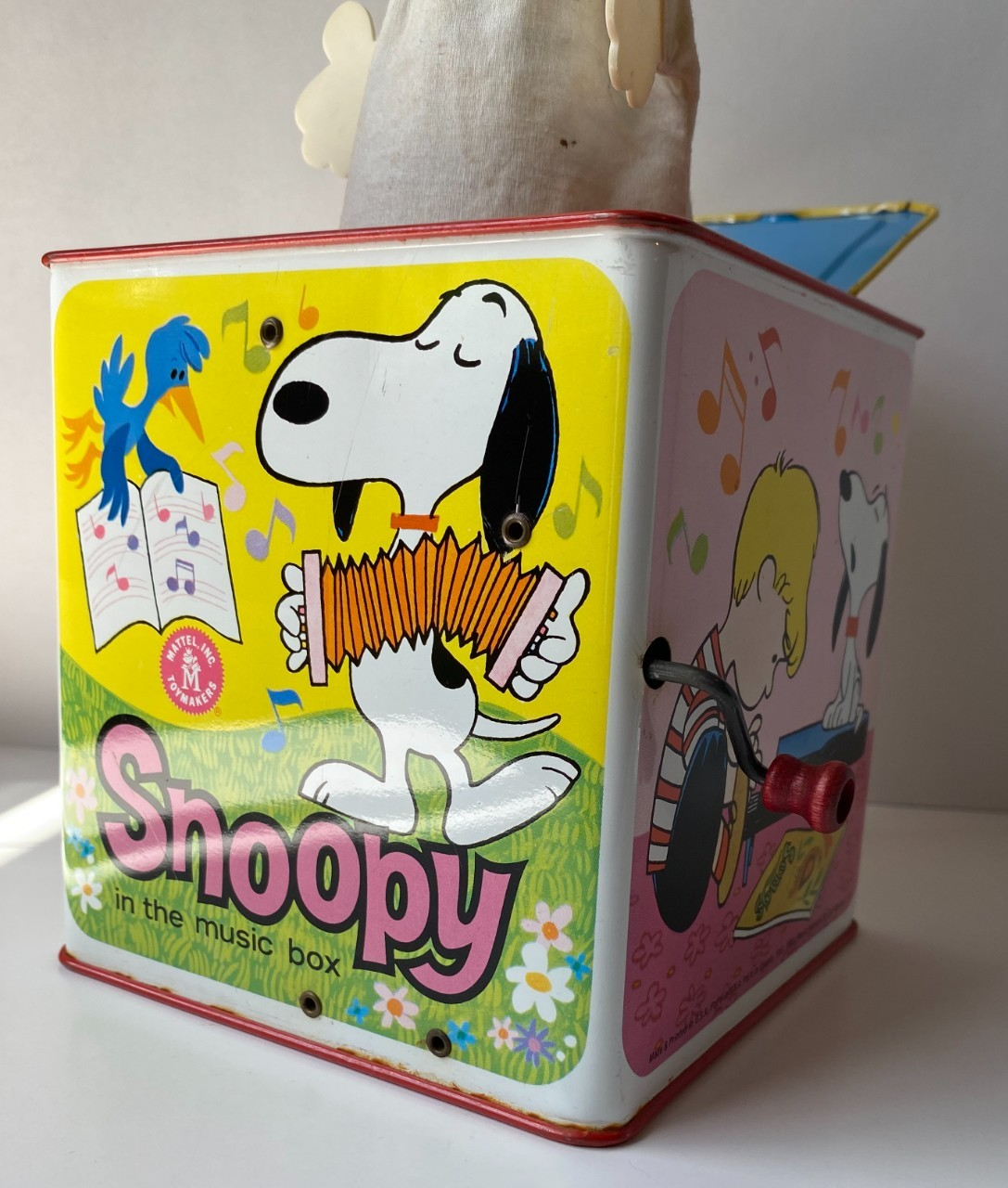 White Snoopy tin lunchbox with red band, blue cap, and text “In the music box” on green background.