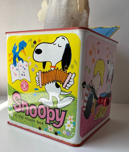White Snoopy tin lunchbox with red band, blue cap, and text “In the music box” on green background.