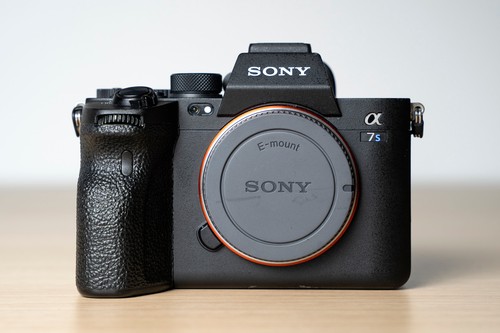 Black Sony Alpha a7S III camera with orange E-mount, textured grip, and two black batteries on a wooden surface.