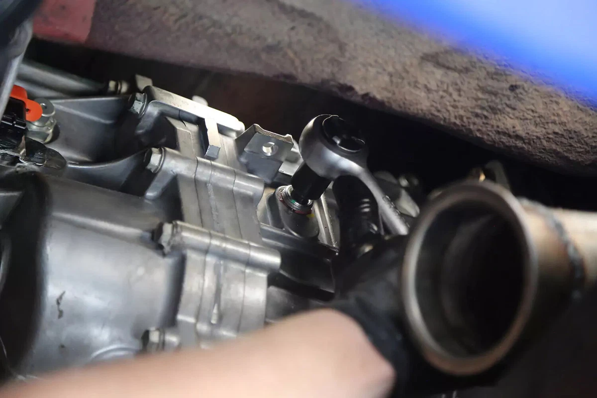 A mechanic wearing a black glove uses a socket wrench to tighten the fill plug of a manual transmission.