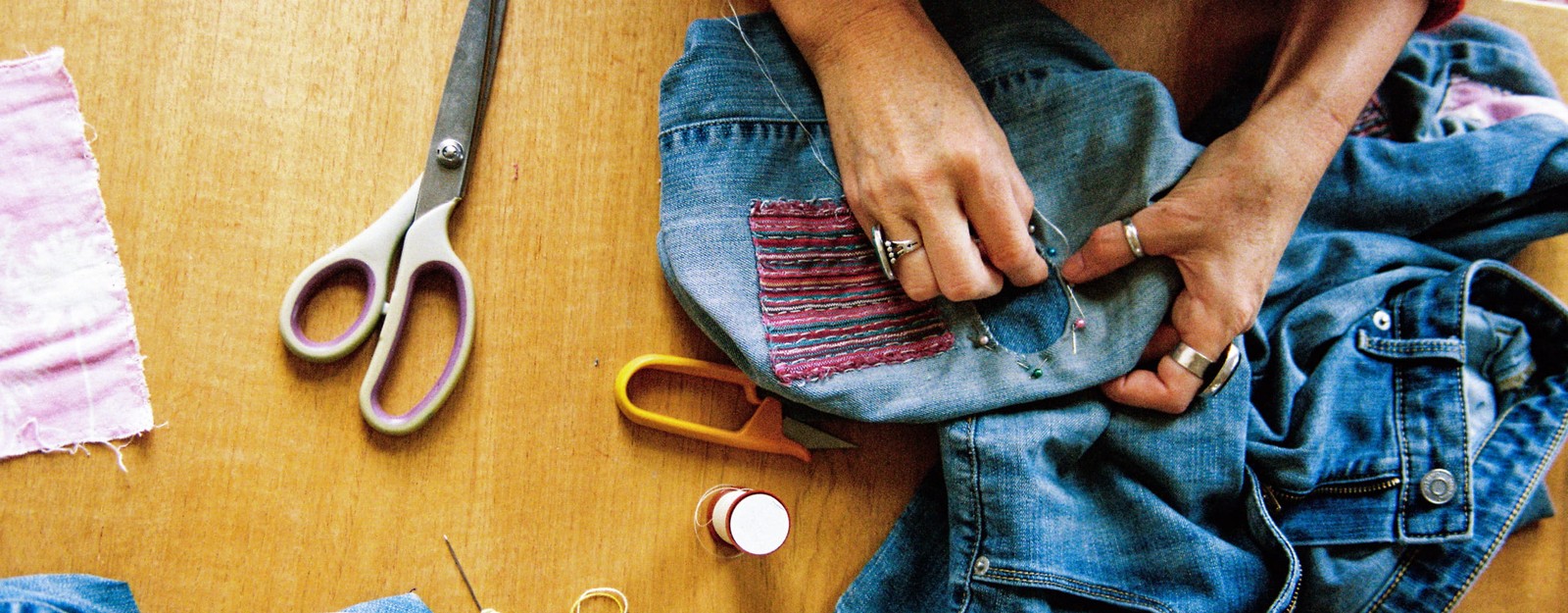Hände nähen mit Stecknadeln einen Aufnäher auf eine blaue Jeans, umgeben von einer Schere, Garn und einem Nadeleinfädler auf einem Holztisch.