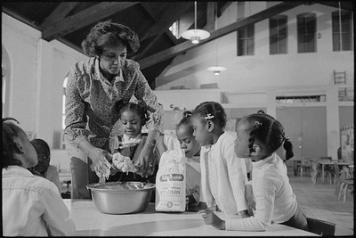 Teacher helping girl put batter in a cake pan, Head Start Program 1979