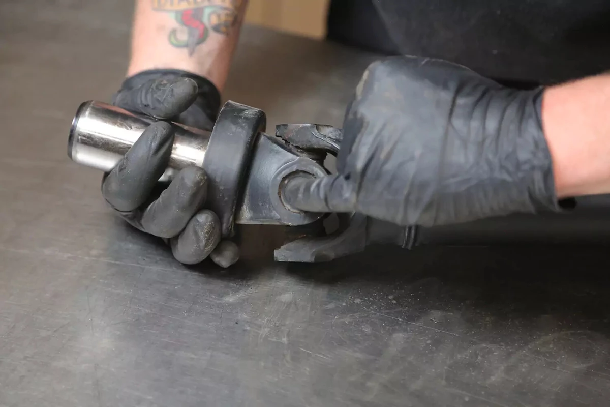 A mechanic wearing black gloves holds a staked U-joint.
