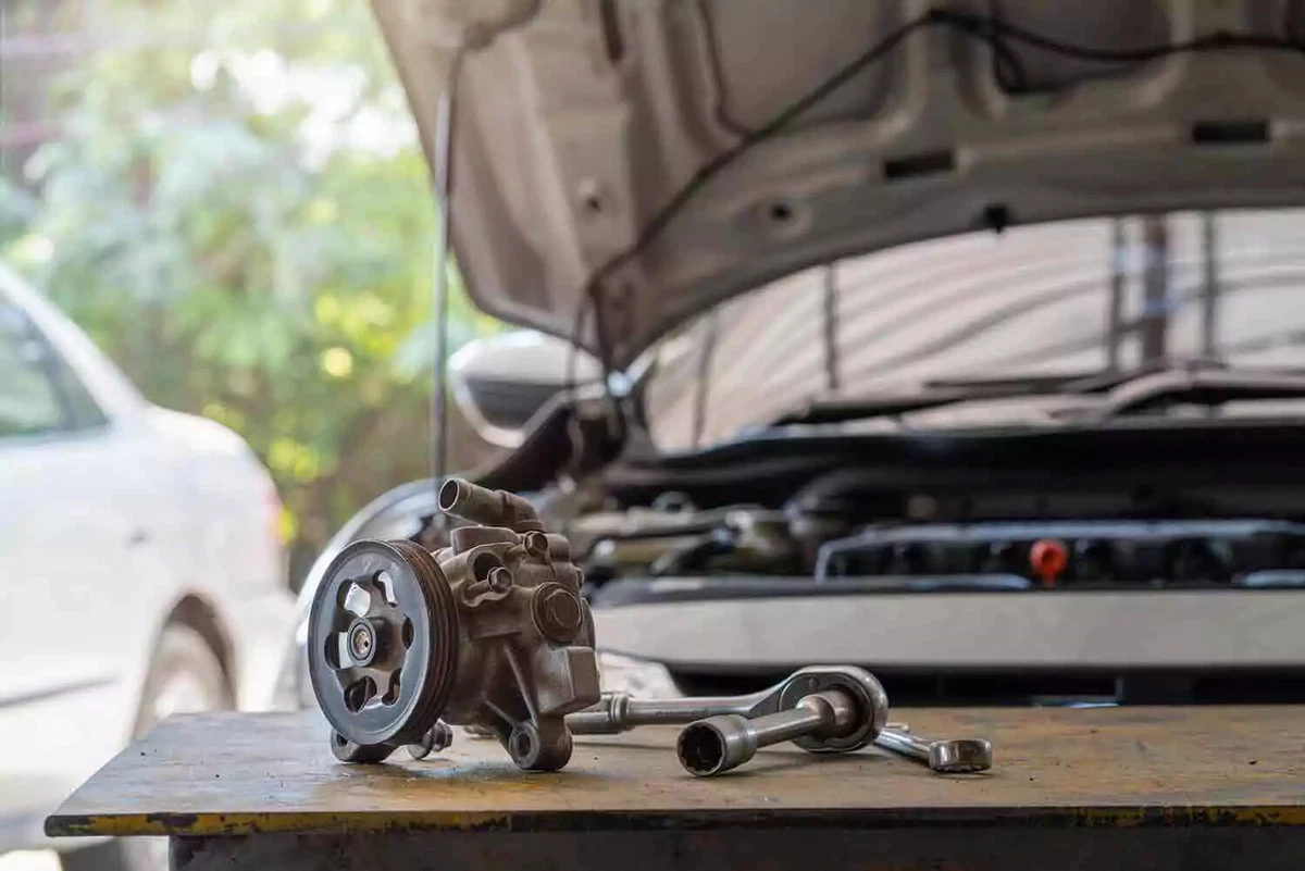 A power steering pump, a ratchet wrench, and a box end wrench on a work table, in front of a car with its hood up.