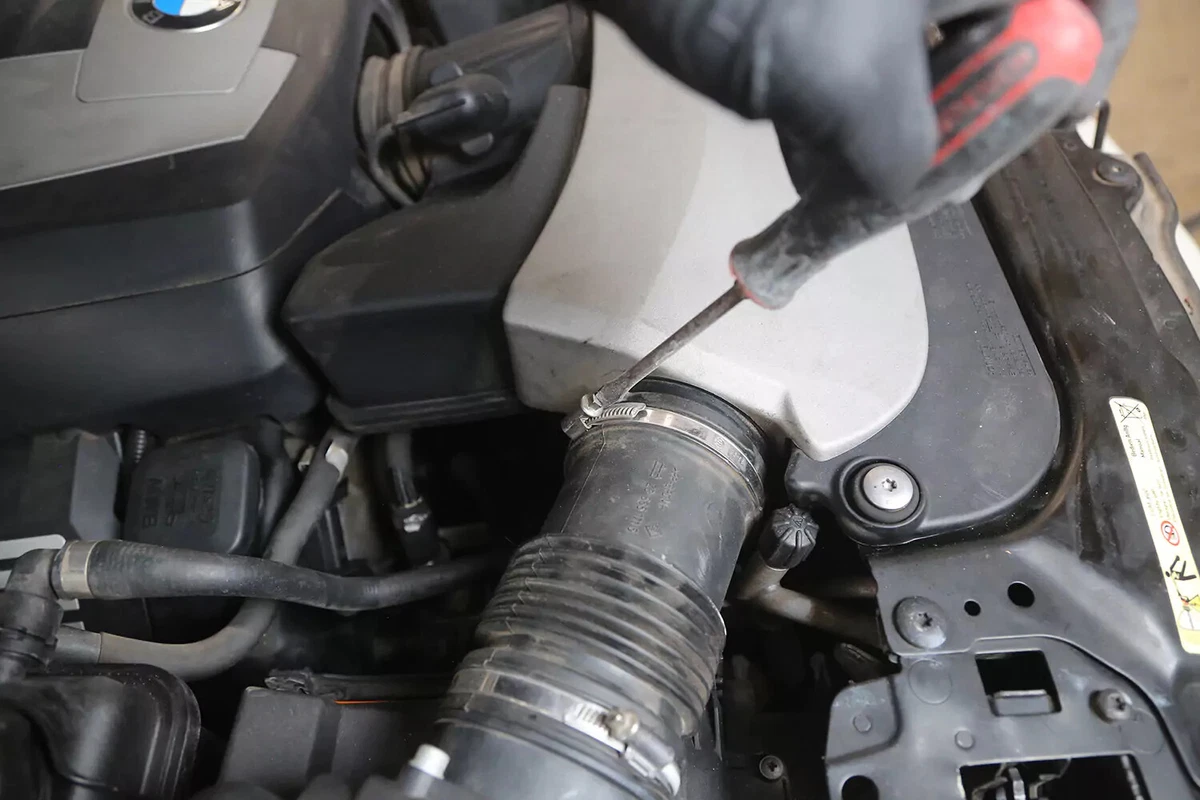 A mechanic wearing a black glove uses a screwdriver to loosen the hose clamp holding the air tubing to the air filter housing.