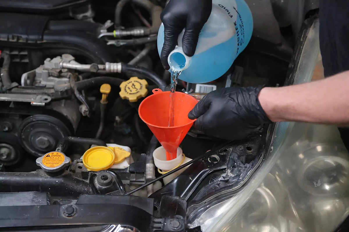 A mechanic wearing black gloves pours windshield fluid through a red funnel into a car's windshield fluid reservoir.