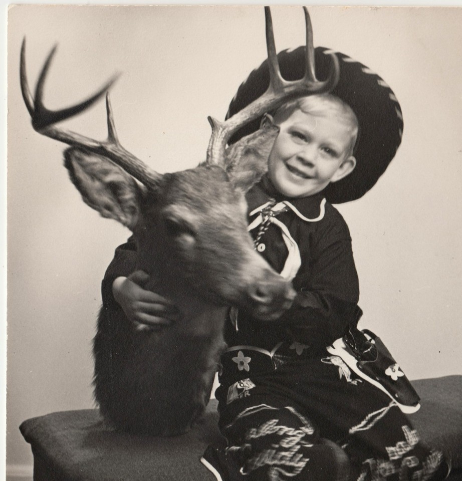 A 1950s snapshot of a cowboy kid posing proudly with a deer head taxidermy — pure Americana weirdness that belongs on a gallery wall.
