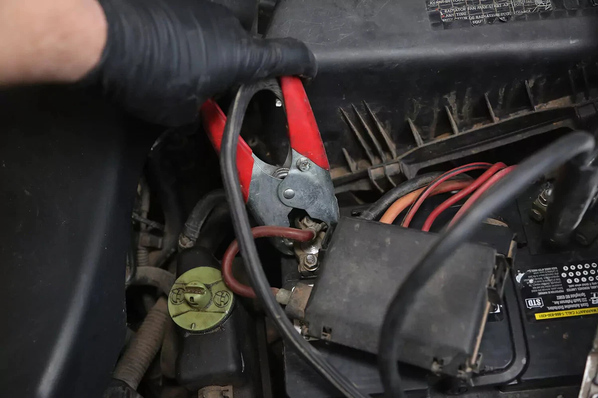 A mechanic wearing a black glove attaches a red handled jumper cable to the postive terminal of a car battery.