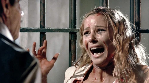 Crying woman in white dress with red marks, locked in cell with man in blue coat.