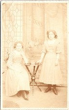 Two Young Women in Long Dresses with Table and Plant, Edwardian Era Postcard