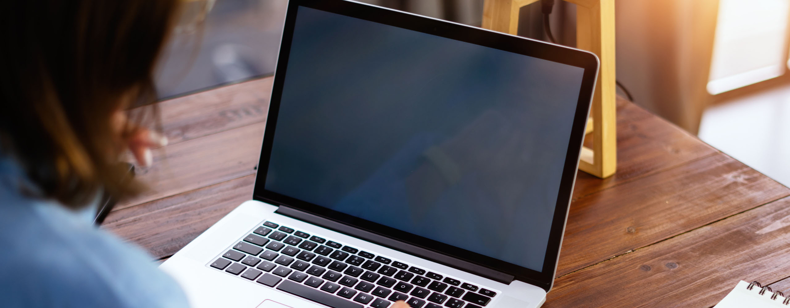 Person arbeitet an einem Laptop mit ausgeschaltetem Bildschirm auf einem Holztisch, umgeben von warmem Tageslicht.