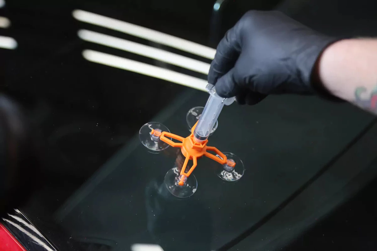 A mechanic wearing a black glove places the syringe from a windshield repair kit into the orange repair jig.