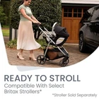 Woman pushing black and silver Britax Willow SC stroller with car seat, brown garage door and green hedge in background.