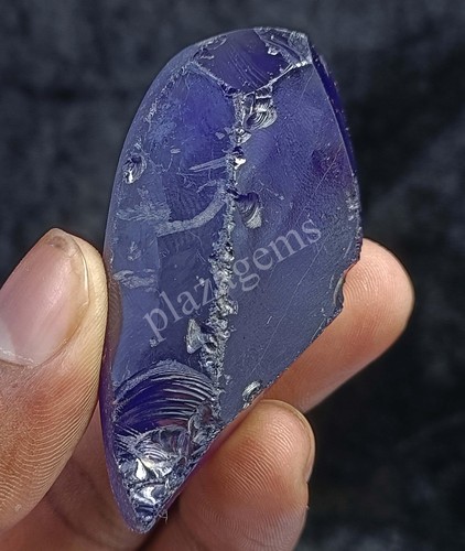 Rough purple zircon 411.05 ct with visible inclusions in metallic blue matrix, held by hand against gray background.