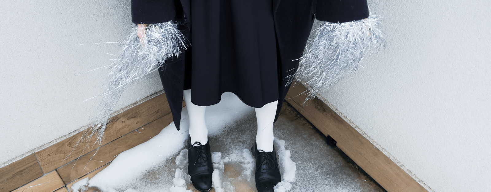 Eine Person mit einem schwarzen Rock, weißen Strumpfhosen, schwarzen Schuhen und einem langen schwarzen Mantel steht im Schnee und hält silbernes Lametta in den Händen.