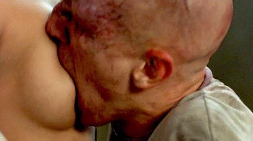 Close-up of a tattooed man's face, red and pale, kissing a woman in prison garb on a wooden surface.