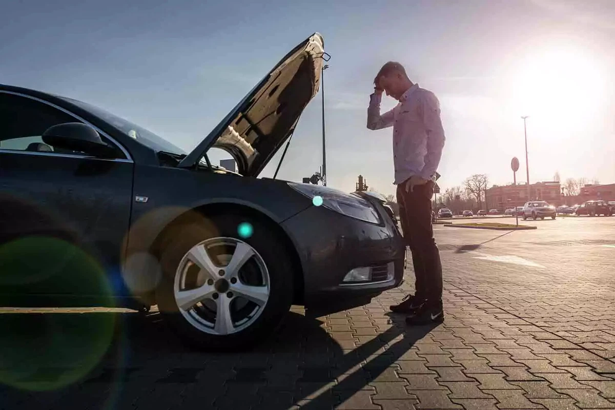 A young man standing next to a broken-down car with its hood up and holding his forehead.