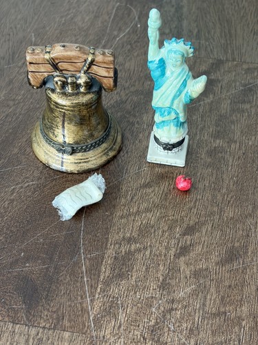 Miniature Liberty Bell with gold accents beside a red, white, and blue Statue of Liberty figure on a wooden surface.