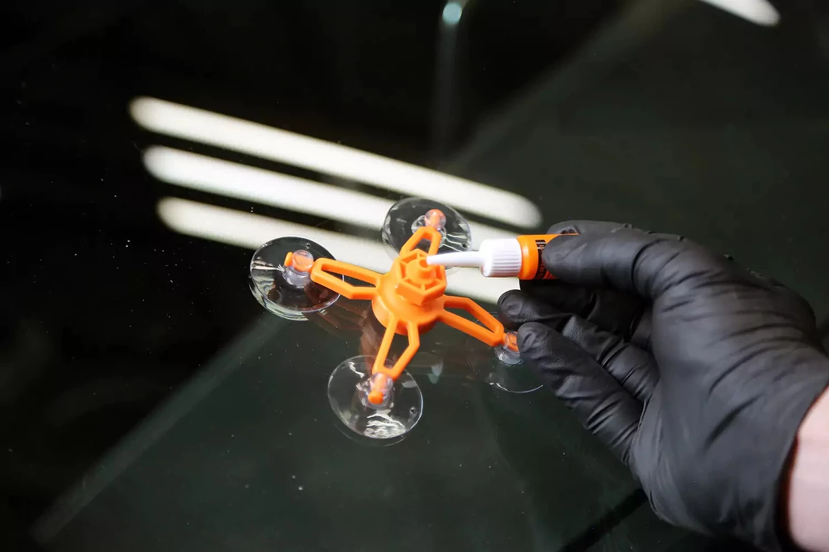 A mechanic wearing a black glove applies repair liquid to the repair jig of a windshield repair kit.