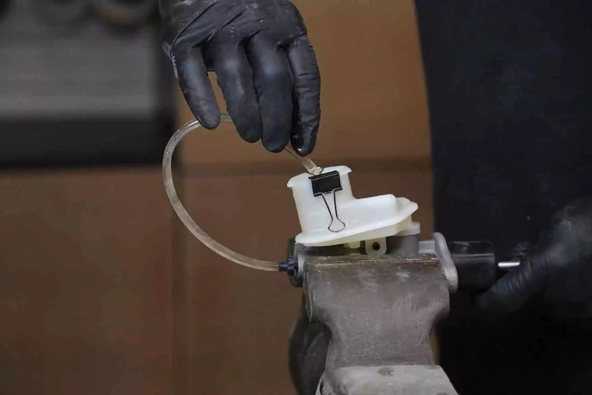 A mechanic wearing black gloves installs a bleeder line in a clutch master and uses a binder clip to hold it in place.