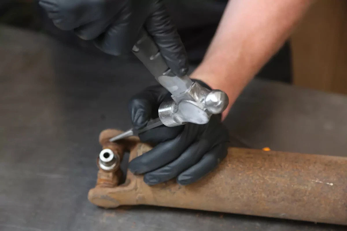 A mechanic wearing black gloves uses a punch and hammer to knock out the inside clip from a driveshaft.