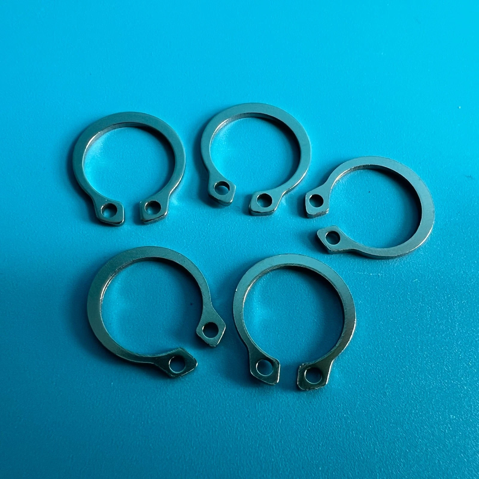 Five silver metal snap C-ring parts arranged in a row on a bright blue background, one with a black text label.
