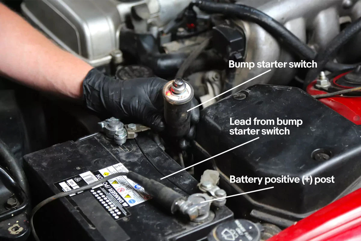 A mechanic wearing a black glove holds a bump starter switch connected to the positive battery post of a vehicle.