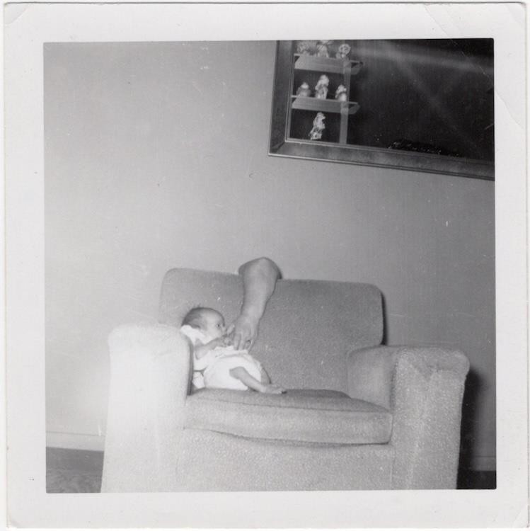 Infant with arm of hidden parent behind chair, 1960s