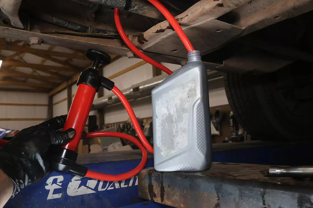 A mechanic wearing a black glove holds a transfer pump with one tube in a bottle of new transfer case fluid.