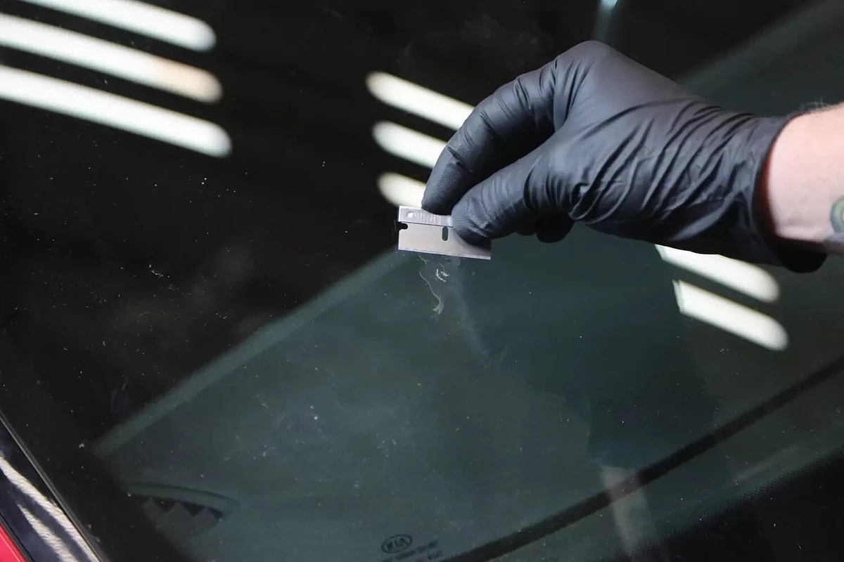 A mechanic wearing a black glove uses a razor blade to scrape away excess material from a repaired windshield.