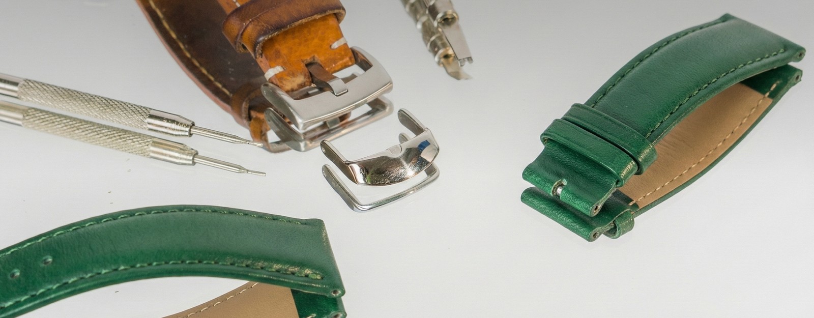 Verschiedene Uhrenarmbänder aus grünem und braunem Leder und mit silbernem Verschluss sowie Uhrenmacherwerkzeuge liegen auf einer hellen Oberfläche.