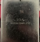 Black metal plate with white etched text reading “U.S.A., 160506 COMPLETE” on a worn, scratched surface.