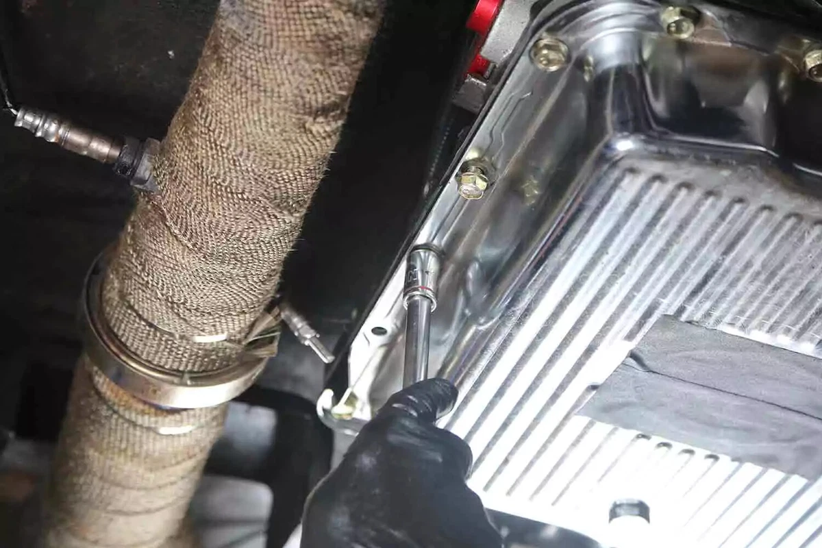 A mechanic wearing a black glove uses a socket and ratchet to tighten the bolts of a vehicle's transmission pan.