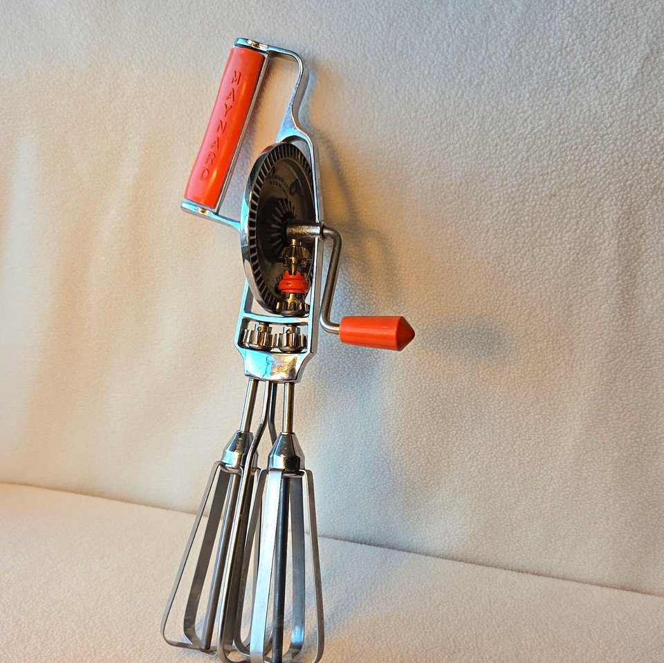 A hand-crank egg beater in salmon orange — the kind of satisfying, zero-electricity tool that makes you feel like you're cooking in a 1940s kitchen.