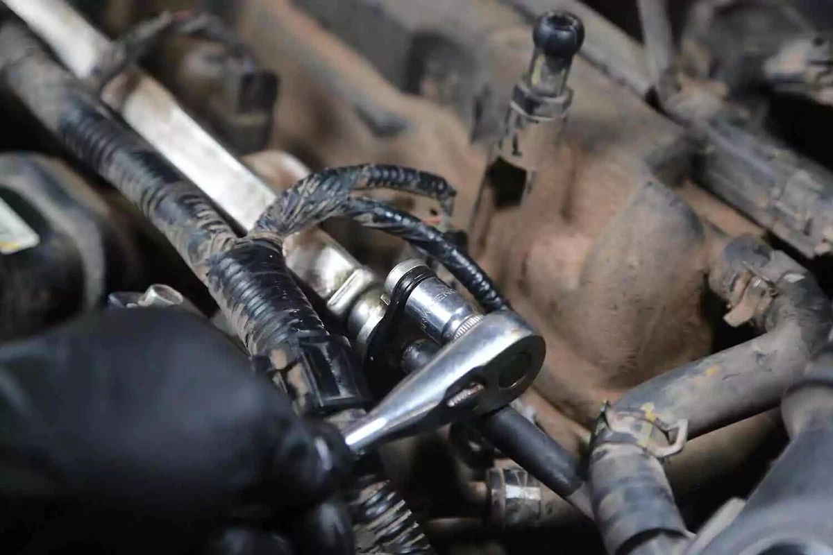 A mechanic wearing a black glove uses a ratchet to reinstall the fuel line.