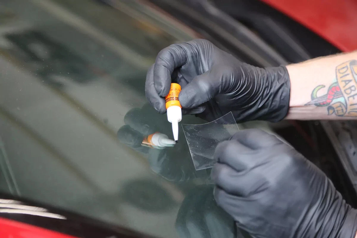 A mechanic wearing black gloves adds a drop of repair liquid to a windshield while holding a clear plastic top slip.