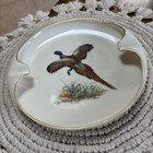 Round porcelain ashtray with gold rim, featuring brown pheasant bird image and gold cigarette rests on woven mat.