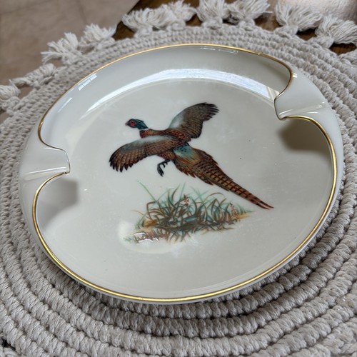 Round porcelain ashtray with gold rim, featuring brown pheasant bird image and gold cigarette rests on woven mat.